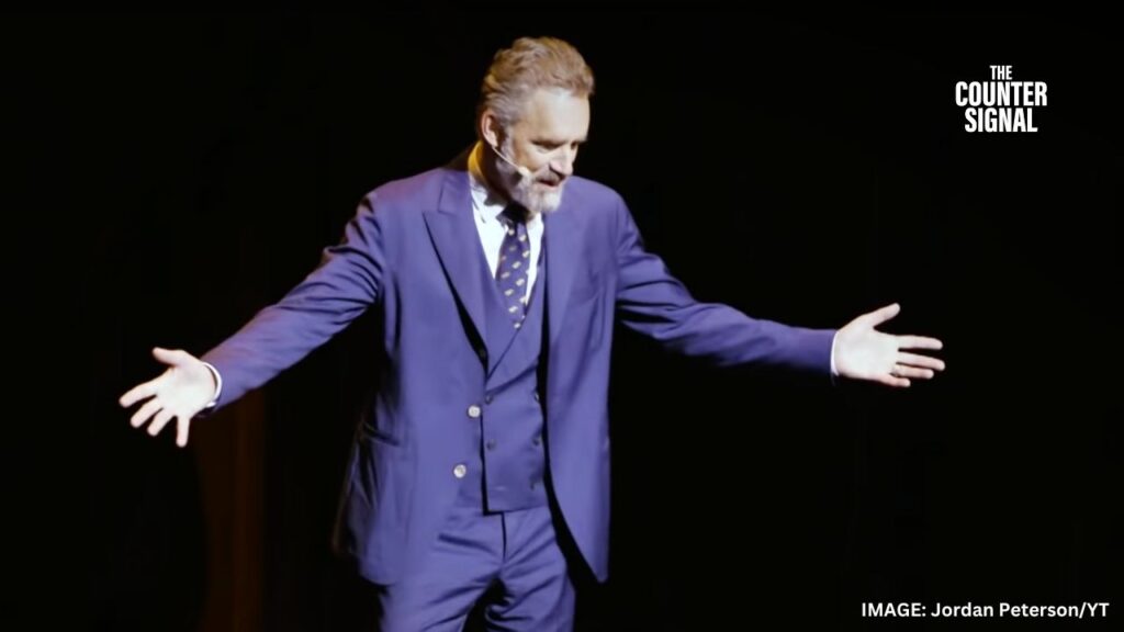 Image of Jordan Peterson with his arms stretched out in a purple suit on stage against a dark background with a slight smile on his face 